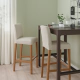 Two BERGMUND bar stools in beige and light wood at dark counter, with cushioned seats and backs, set against light green wall and light wooden floor.