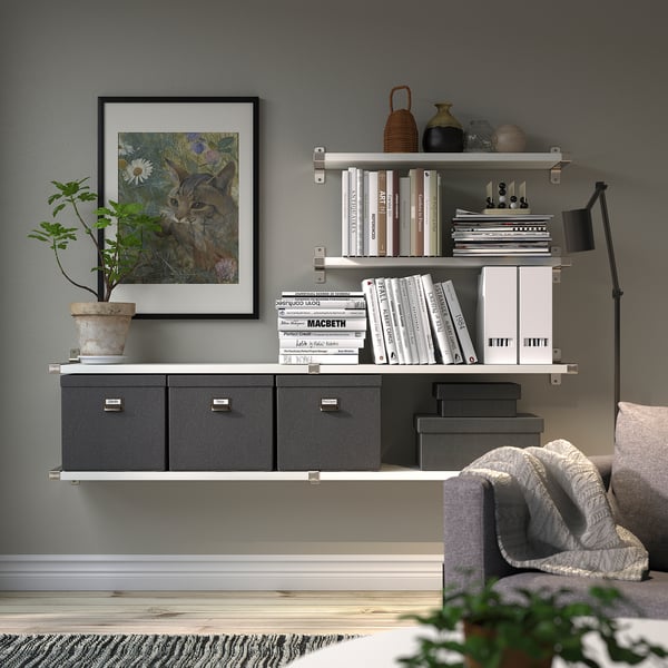 Modern living space with wall shelf displaying books and decor, including cat art, plant, and lamp. Storage boxes below and sofa with blanket.