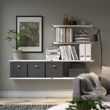 Modern living space with wall shelf displaying books and decor, including cat art, plant, and lamp. Storage boxes below and sofa with blanket.