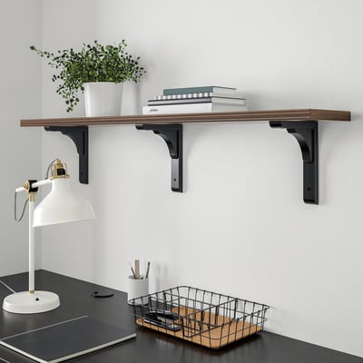 BERGSHULT wooden shelf, walnut-patterned, wide & low, holds stack of books, plant, and lamp. Metal brackets. Black desk below with basket and lamp.