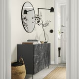 Contemporary entryway grey BESTÅ storage unit with books, glass vase, and wicker basket under large round mirror with sleek black lighting.