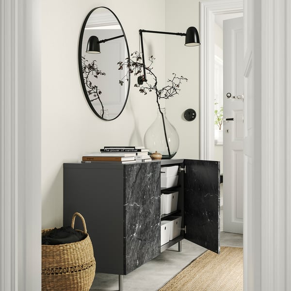 Modern entryway with black BESTÅ storage, white boxes, books, vase, mirror, & wall sconces.