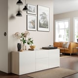 White BESTÅ sideboard holds items like vases and books, set under hanging frames. Its next to an orange sofa and large window.
