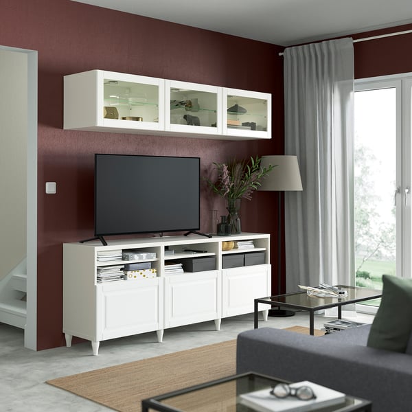 Modern living room with BESTÅ tv stand, flat screen, open shelves, dark maroon wall, grey sofa, glass coffee table, soft lighting.