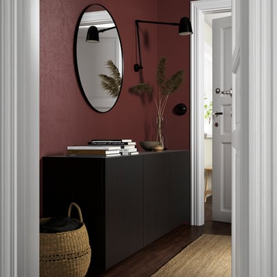 Dark BESTÅ cabinet holds books and vase, mirror above, basket beside, red wall.