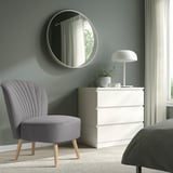 Cozy bedroom corner with grey BILLHAMN chair, white dresser, plant, & round mirror. Soft and comfortable, fits neatly.