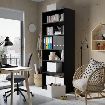 Black BILLY bookcase with books and storage, taller than desk, against walls with windows.