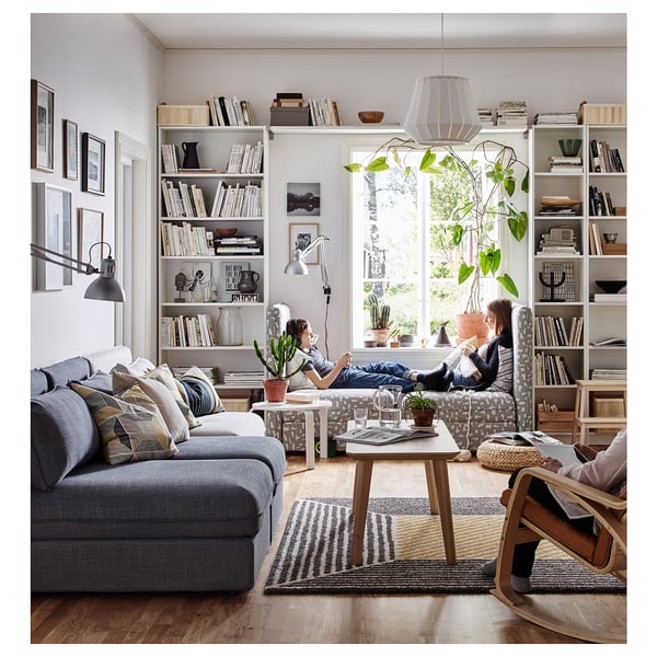 Modern living room with gray sectional, wooden tables, striped rug, bookcases, hanging lamp, white walls, and windows with greenery views.