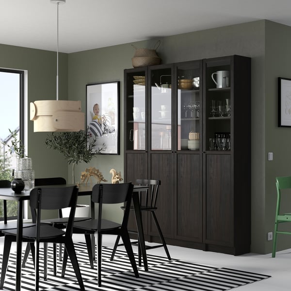 Modern dining room with a tall black BILLY bookcase filled with glassware. It stands next to a black table and chairs with a large window nearby.