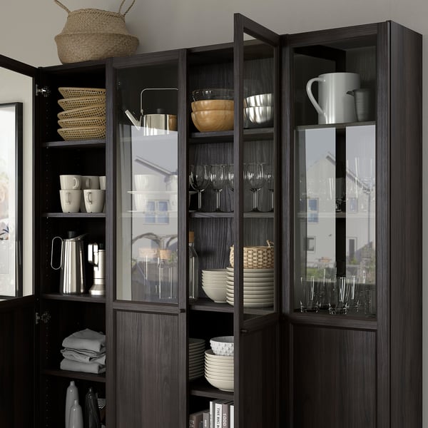 Dark wood BILLY bookcase with glass doors displaying dishes, cups, and glasses neatly organized on multiple shelves.