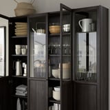 Dark wood BILLY bookcase with glass doors displaying dishes, cups, and glasses neatly organized on multiple shelves.