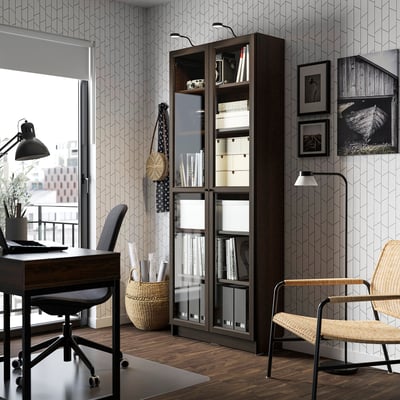 A home office with a tall BILLY bookcase, filled with books, storage, and a wicker chair by a large window.