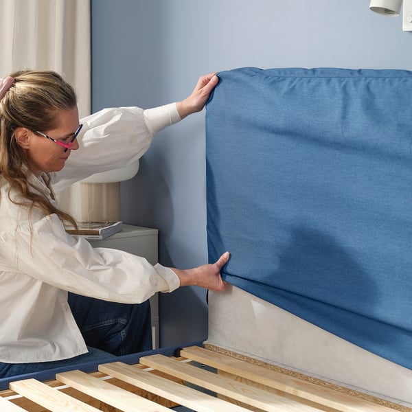Image description: a person fitting a blue headboard cover on a BLÅFJÄLLET bed frame.