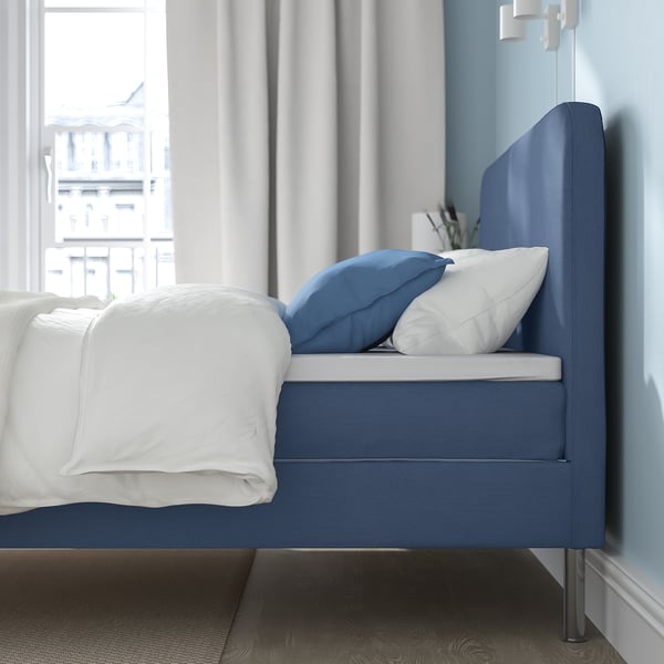 Modern blue bed in a room with white curtains. The bed has a clean, minimalist design and includes white and blue pillows.