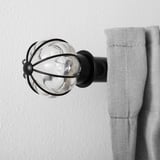 Glass and metal curtain finial mounted on wall, holding light gray curtain.