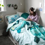 Girl in purple hoodie reads on wave-patterned bed with headphones and green pillow. Nightlight on wall.
