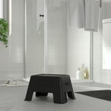 Modern bathroom with black BOLMEN step stool, smaller than slippers, beside glass shower. White tiles, gray mat, bottles, and towel.