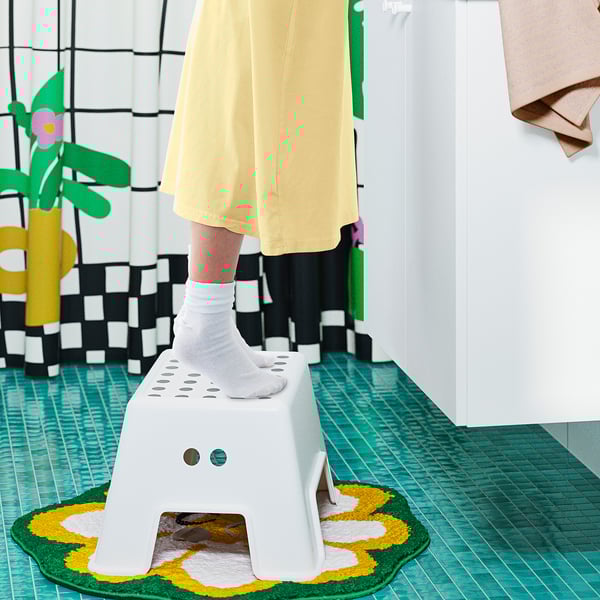 Kid stepping on small white BOLMEN stool, yellow dress, green rug, colorful decor.