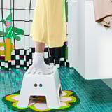 Kid stepping on small white BOLMEN stool, yellow dress, green rug, colorful decor.