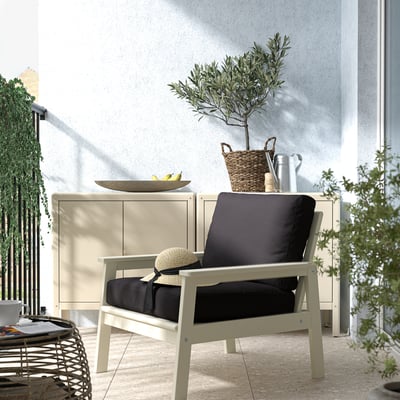 Modern balcony with cream armchair, black cushions, and side table. Basket with plant and decor items. Surrounded by greenery.