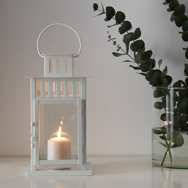 Lit lantern BORRBY on white surface, warm glow, minimalist design, eucalyptus in clear vase.