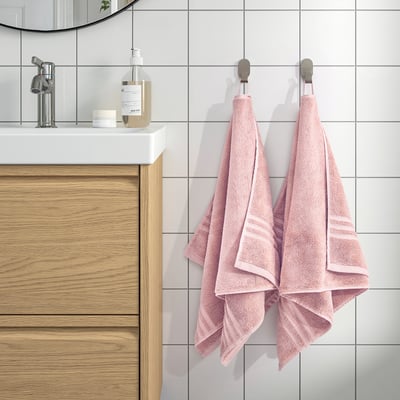 Two pink BROKGLIM towels hang on wall hooks beside a wooden vanity with a round mirror, white countertop, and soap dispenser.