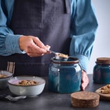 Person scoops granola from a blue BRUGDHAJ jar into a bowl. Table displays other jars, bowls, and utensils.