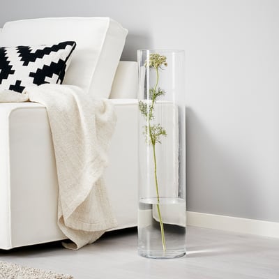 Tall clear CYLINDER vase with single long-stemmed plant beside white armchair.