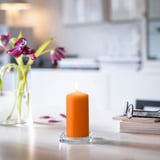 Small orange candle in glass holder beside pink flowers and books on white table.