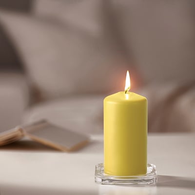 A lit yellow DAGLIGEN pillar candle on a clear glass base, larger than nearby matches, on a table.