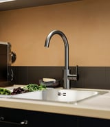 Modern kitchen sink with DELSJÖN pewter faucet, vegetables, and bowl against brown backsplash.