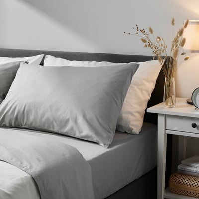 The DVALA light grey cotton pillowcase on a bed with a grey headboard. Taller than a bedside clock, shorter than a vase with dried flowers.
