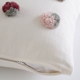 A beige cushion cover with pink and gray pompoms, featuring a hidden zipper on the side.