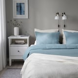 Cozy bedroom with dual lamps, vase, books, and glass. Blue bed, 2 pillows, warm blanket, art, curtain. Bed larger than nightstand.