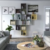 Modern living room with gray EKET wall-mounted shelves displaying books, vases, and art. Sofas, coffee table, and plants complete the space.