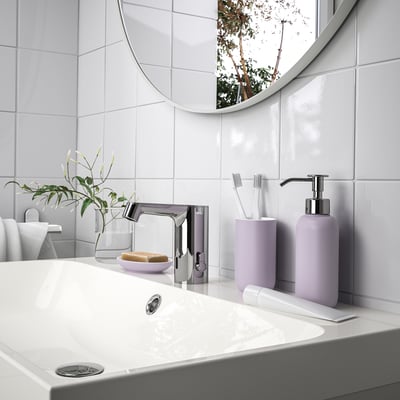 Modern white bathroom with gray counter and sink. Large round mirror above. Purple toiletries in lavender holder. Plant in clear vase.
