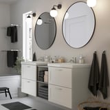 Modern white bathroom vanity with two sinks, large mirrors, and open shelving. Black towels hang on either side. Circular light fixtures above.