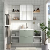Modern bathroom with green ENHET vanity, mirror, open shelves, plant, towels, toiletries, and sink under window.