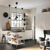 Modern kitchen with white high cabinet, black counter, stools, and hanging lamps, open shelves with greenery, black and white decor.
