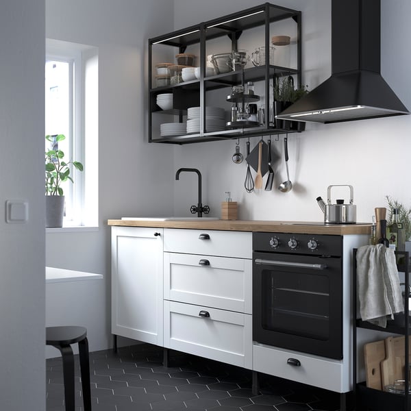 Modern kitchen with white & black ENHET cabinets, butcher block counter, black hood, and dishes on open shelves. Green plant by window.