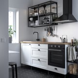 Modern kitchen with white & black ENHET cabinets, butcher block counter, black hood, and dishes on open shelves. Green plant by window.