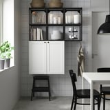 Modern kitchen with ENHET white cabinets, black open shelves, stacked dishes, and books. A black chair and stool are nearby.