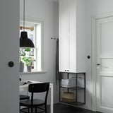 Minimalist workspace: ENHET white cabinet, tall & narrow, holding books & basket. Black desk & chair, with black lamp. Door & windows beside.