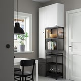 Modern room with white cabinet, black cart, and chairs. Cabinet has storage and glass section. Cart holds bottles and books. Window with plant.
