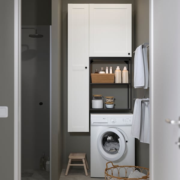 Modern bathroom with white ENHET cabinets, washing machine, open shelves with laundry supplies, towels, and a black shower in the background.