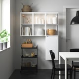 Modern kitchen corner with ENHET white storage, black cart, and black chairs around a white table. A plant by the window.