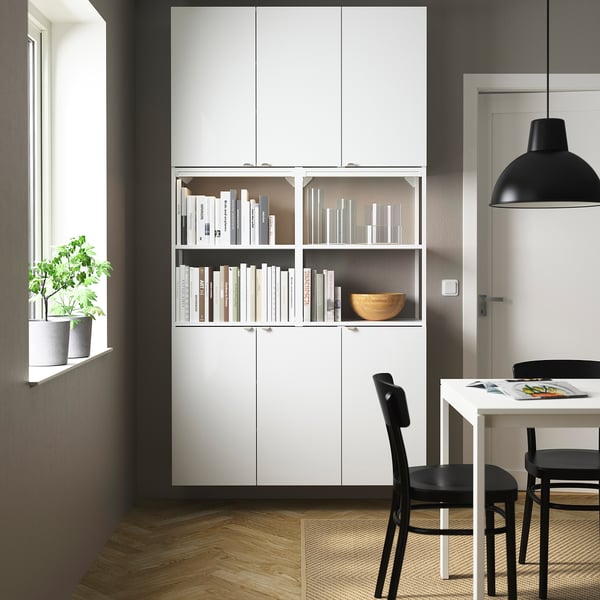 Minimalist room with ENHET white cabinet. Books and decor displayed, potted plant by window, black chairs around table. Black pendant light.