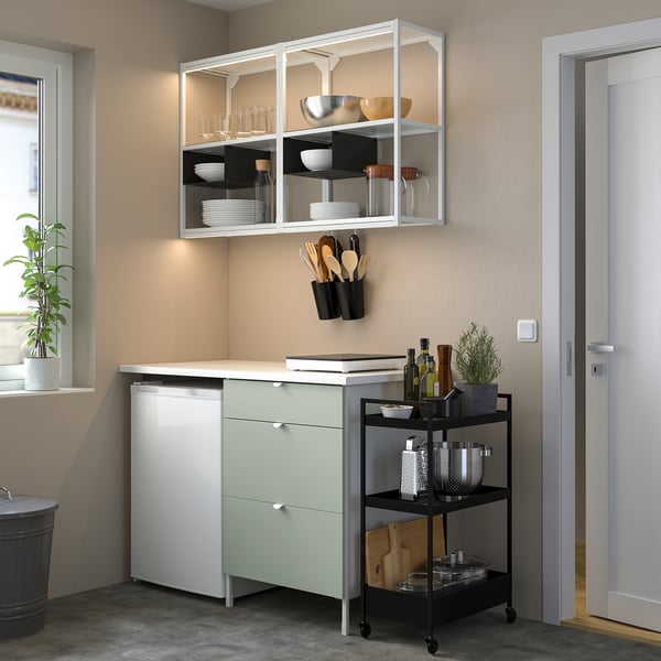 Small kitchen setup featuring ENHET pine green cabinet with drawers, metal shelves, utensils, bottles, and plant.