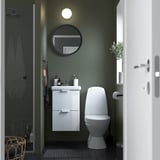 Modern bathroom with ENHET white cabinet, round mirror, toilet, and shower. Green walls and sleek design.