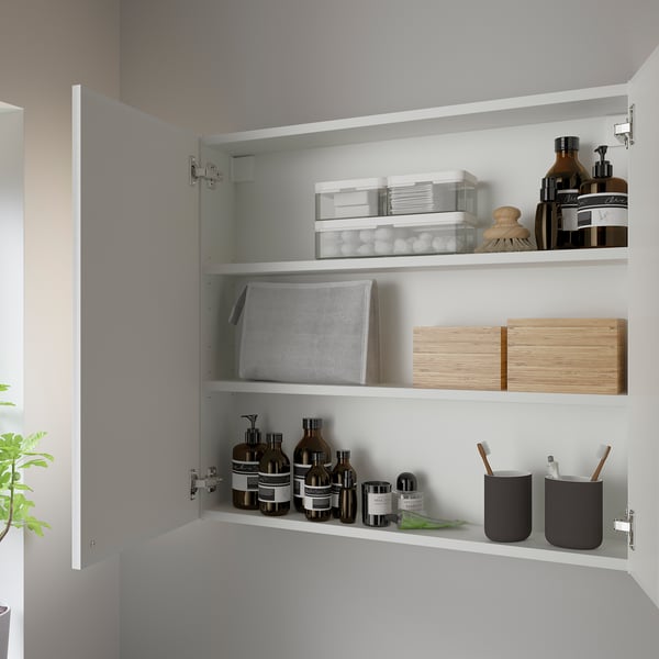 Open white bathroom cabinet with three shelves storing toiletries, jars, and containers for organized accessible storage.